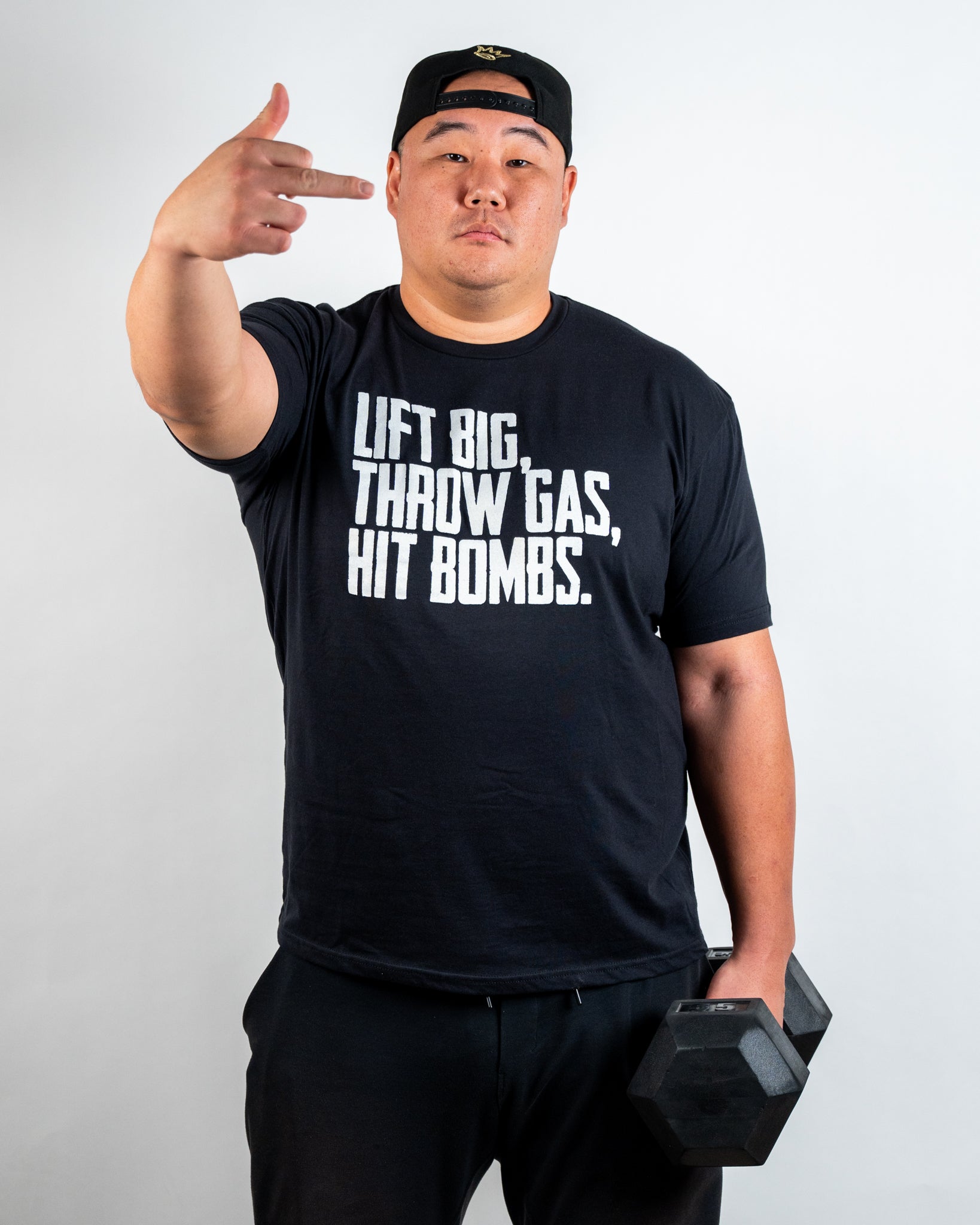 Model in black Juco Bandit T-shirt holding a dumbbell, showcasing 'Lift Big, Throw Gas, Hit Bombs' slogan.